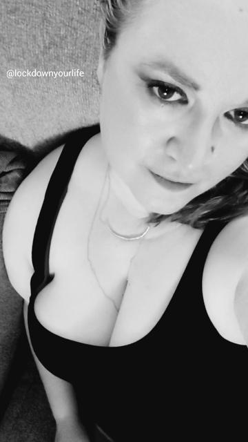 A black and white photo of a woman with winged eyeliner and wearing a sleeveless black shirt, smirking at the camera. She has on a layered necklace and the camera is angled down toward her face and shoulders. She has really nice bewbs.