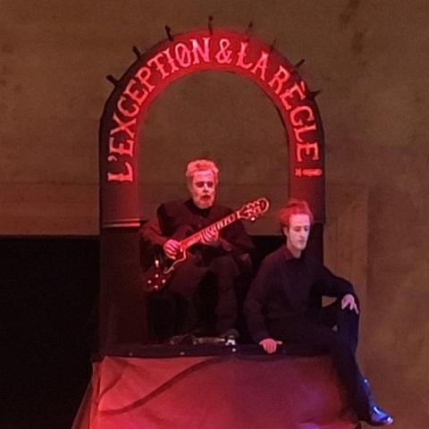 photo de deu personnage assis en hauteur.
celui de droite à une main sur les genoux croisés.
celui de gauche joue de la guitare électrique.
ils portent des vêtements noirs et du maquillage blanc sur le visage.
au dessus d'eux le titre du spectacle en arc de cercle.
La lumière d'ambiance est rouge.