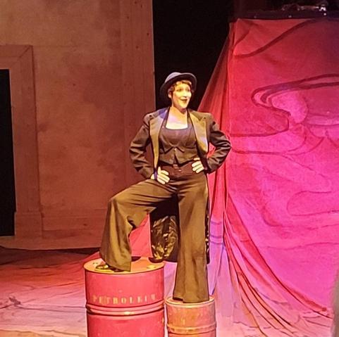 Montée sur deux bidons de pétroles formant un escalier, une comédienne vêtue de noir fait un grand sourire les mains sur les hanches.
La photo est également dans des teintes rouges.