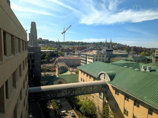 View through a window along a tall wide building that's to the left and low buildings and roof to the right. A tall tower is in the distance as is a very tall crane