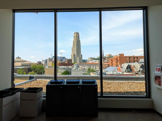 A 3 panelled window that fills the whole side/wall with a bit of gravel outside and the cathedral of learning tower framed in the middle window. Blue skies and thin wispy clouds