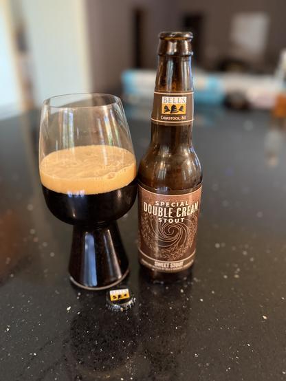 A stout glass of dark beer with a tan head. The brown bottle has a label on shades of brown
