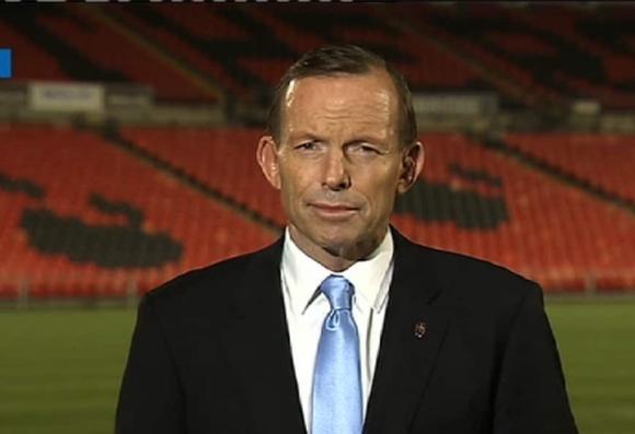 Tony Abbott stands in front of empty stadium seats, looking serious. The background features orange seats with dark patterns.