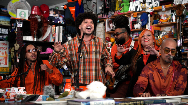 A group of people performing in the Tiny Desk studio — rapper PJ Sin Suela and his band and sign-language interpreter. Sin Suela, in a plaid shirt, is smiling, The shelves behind him and his bandmates are crammed with books and memorabilia.