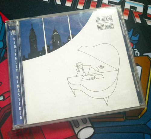 Front cover of Joe Jackson's "Night And Day" on CD. A minimalist line drawing of the singer at his piano appears on the lower right corner. In the upper left the New York City skyline at night is visible through three windows behind him.
