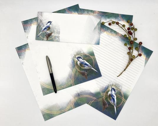 Product photograph of several sheets of illustrated bird stationery featuring a Black-Capped Chickadee against a pastel textured edges of teal and greens with light glowing circular motifs floating throughout.