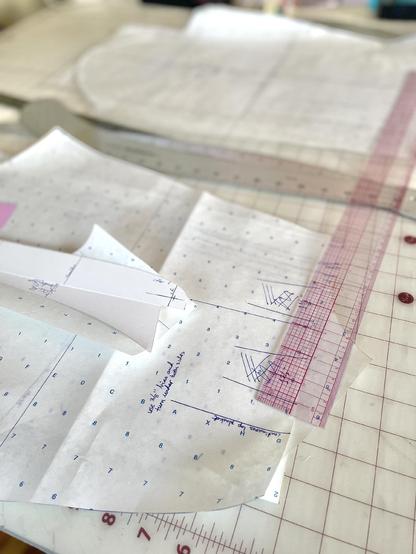 A pile of pattern pieces for a button up shirt is held down by a clear ruler with red numbering. In the background in another pile of pattern pieces for a bodice sloper. All pieces are dragged on semi-transparent white pattern paper with blue grid marks.