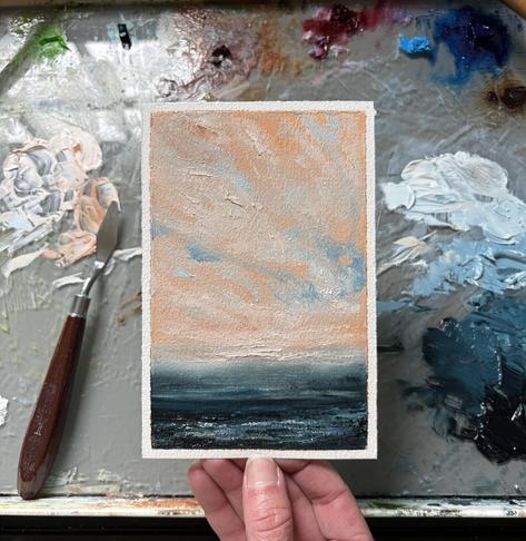 Photo of a hand holding an original seascape oil painting by Tisha Mark, "Peaceful Spring Sunset" 6"x4" oil on Arches oil paper (2025). A seascape painting with a textured orange-toned sunset sky over a deep blue sea. A paint palette with puddles of oil paint and a palette knife are visible in the background.