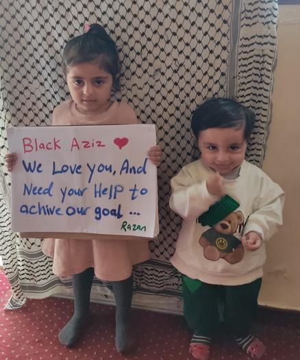 Image shows two small children, one child has a sign which says "Blacm Aziz, as love you, and need your help to achieve our goal."