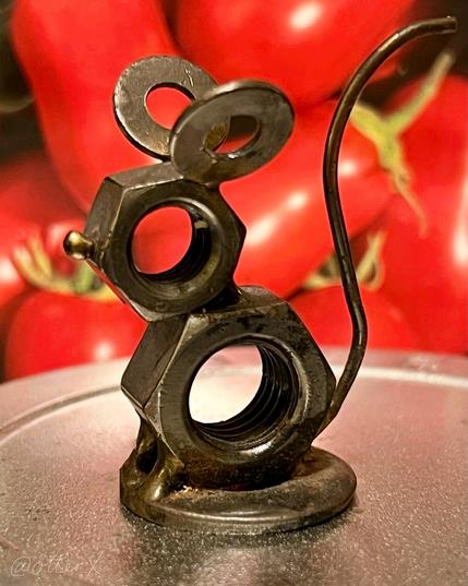 The little life-size mouse made of metal nuts and washers is placed on top of the silver tin in front of the packet of Roma tomato seeds, which depicts some plump red oval tomatoes.