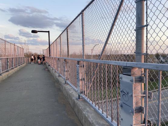 Looking along a pedestrian bridge with metal fence structures on both sides. The metal is pink in the setting sun, with blue shadows. At the end of the bridge before the down ramp there's a group of kids hanging out.