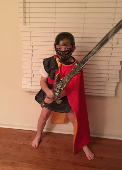 Image shows a little boy dressed up in chain mail, wearing a helmet, red cape and brandishing a big sword. He is pant less and barefoot.