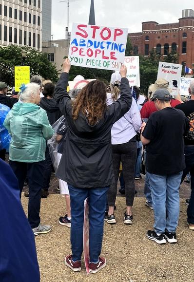 Woman holds up hand lettered sign: "STOP ABDUCTING PEOPLE YOU PIECE OF SHIT"
