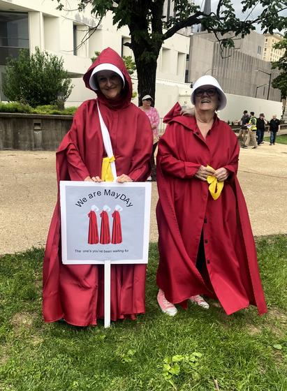Two women dressed as Handmaidens. Sign: Three stylized handmaidens in profile. Caption: "We are May Day / The one's you've been waiting for"