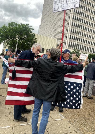 Woman in suit jacket and jeans, with her back to the camera, holds a large upside down American flag. An elderly man leans toward her smiling.