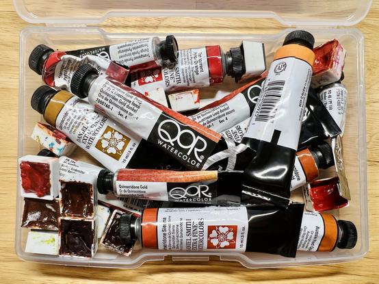 A small plastic container filled with various tubes of paint in all different shades of orange. There are also a bunch of half pans, filled with dried paint, mixed in to the chaos.