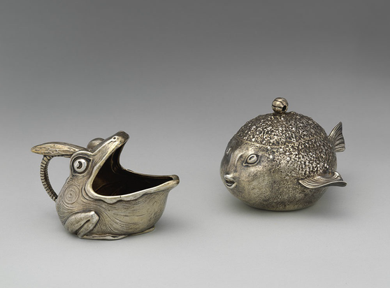 official museum photo of the two objects side by side facing each other in side profile on gradient grey background silver creamer shaped like an open-mouthed frog (left) and sugar bowl shaped liked a puffed-up pufferfish (right)