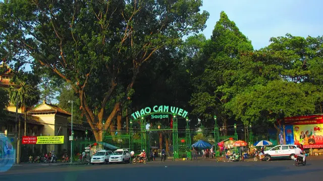 The image features a large green gate that reads "Thảo Cầm Viên Saigon," which translates to "Saigon Zoo and Botanical Gardens." Surrounding the entrance are tall trees and some vehicles, alongside a few people and stalls, suggesting an outdoor area likely associated with leisure and recreation. The atmosphere appears vibrant, indicative of a lively urban park setting.

Image Credits: Wikimedia / trungydang / CC BY 3.0
