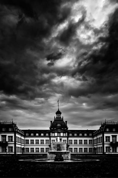 Black and white. Portrait format. A castle with a fountain in the opulent front garden under a dramatically contrasting sky.