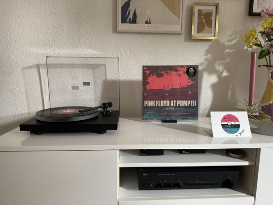 A record player on the left playing a black vinyl record. In the middle, the red green cover. On the right, a painted card.