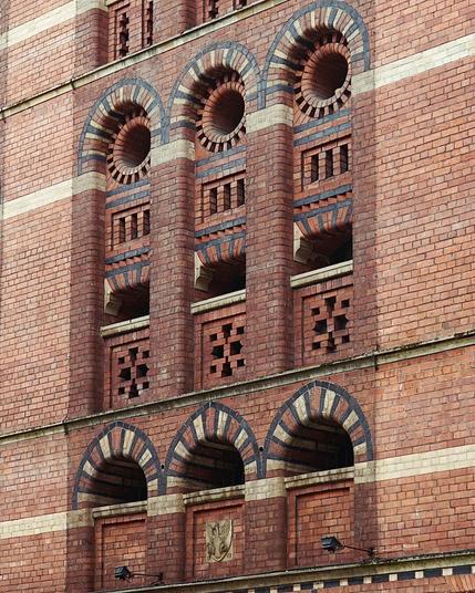 Ouvertures d’inspiration byzantine dans une façade en brique ancienne. Elles sont groupées par trois verticalement. Une alternance de briques rouges, claires et noires souligne le bord des ouvertures en plein cintre (en haut) ou en ogive (en bas).