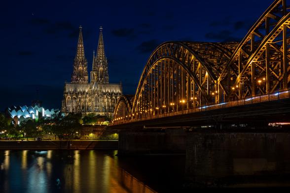 Das Bild zeigt eine Nachtaufnahme der Hohenzollernbrücke und des Kölner Doms von der gegenüberligenden Rheinseite.