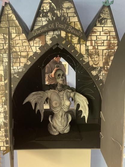 a sculpture of a church-like house with three naves and a large, now open, double-leaf gate. The façade of the house is covered with small tile-like picture cut-outs showing various female film stars from the 1930s to 1960s. Above the gate is a sign that reads 'Shrine of past Beauty'. A female sculpture with bat-like wings is placed in the open shrine in front of a mirrored surface. She is naked and partially skeletonized, just like her skull