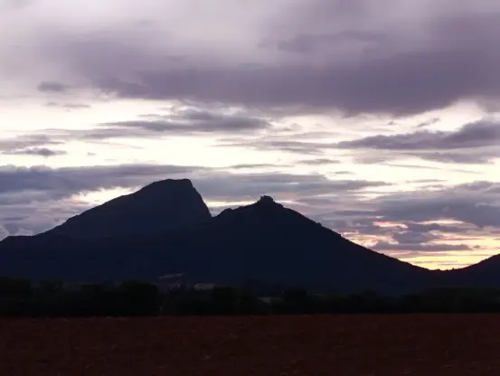 Un paysage de fin de journée, avec un premier plan constitué d’un champ de terre nue et sombre. Derrière ce champ, une bande de végétation arbustive sépare le sol cultivé des reliefs montagneux. Deux sommets se détachent sur l’horizon : l’un, plus massif, à gauche, le Pic Saint Loup, et l’autre, plus effilé, à droite, avec un petit château en ruine à peine visible au sommet du second. Le ciel est largement couvert de nuages.