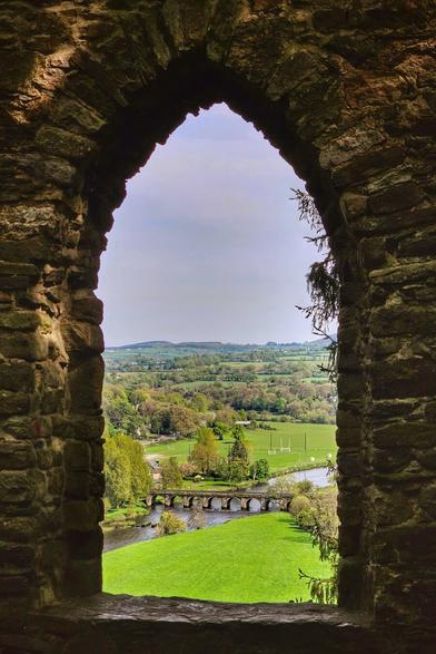 A view of the green Irish countryside, a river, and a stone bridge seen through an arched stone window.