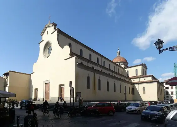 The image depicts a large building that resembles a church or cathedral, characterized by its yellow facade, round windows, and a dome. The architecture appears to reflect Italian styles, and there are vehicles parked in front of it, along with some people. The sky is blue with some clouds, suggesting it was taken during the day.

Image Credits: Wikimedia / Lucarelli / CC BY-SA 3.0