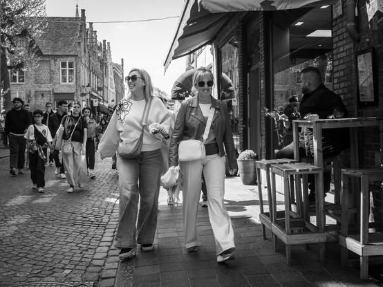 The image depicts a lively street scene in Bruges, Belgium. The photograph is in black and white, emphasizing the textures and contrasts of the scene. In the foreground, two women walk side by side, both wearing sunglasses and carrying bags. The woman on the left wears a white sweater with a floral design and jeans, while the woman on the right sports a leather jacket and white pants. They are walking on a cobblestone street, with a brick building featuring large windows and an awning in the background. The street is bustling with pedestrians, some of whom are carrying shopping bags, suggesting a popular shopping area. The architecture of the buildings, with their gabled roofs and brick facades, adds to the historic charm of the scene.