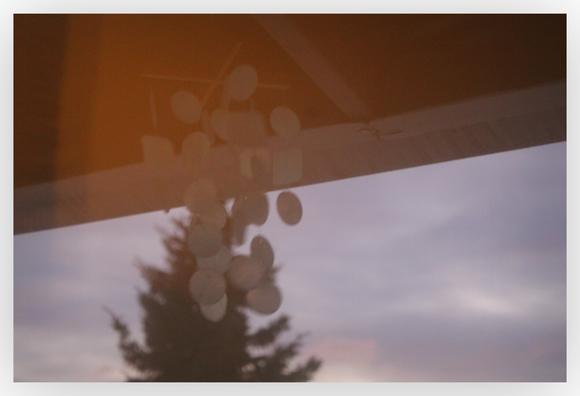 The Sun covering the windows in orange light. through the windows are wind chimes, just under a roof awning. Grey clouds behind.