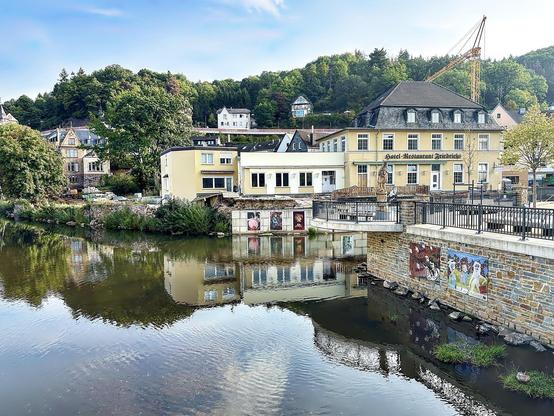 This image depicts a scenic riverside view featuring a mix of commercial and residential buildings. The central structure is labeled as "Hotel-Restaurant Rheinblick," suggesting it's a hotel with a restaurant that probably offers views of the river. The environment is peaceful with lush greenery in the background, including trees and hillside homes that add to the charming setting. There's a calm river in the foreground that beautifully reflects the buildings and the sky, enhancing the serene and picturesque quality of the scene. The architecture and the layout indicate a European setting, possibly in a small town known for its hospitality and beautiful landscapes.