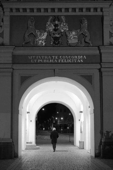 Ein Mann läuft nachts durch einen erleuchteten Torbogen. Der Mann läuft mit dem Rücken zum Betrachter. Beide sind mittig zum Torbogen ausgerichtet. Auf der Fassade ist eine Inschrift angebracht: „Sit intra te concordia et publica felicitas“ – In deinen Mauern herrsche Eintracht und öffentliches Wohlergehen. Über der Inschrift flankieren zwei Löwen drei Wappen. Durch das Tor hindurch sind leere Straßen mit Ampeln und Straßenlaternen im Hintergrund erkennbar.

A man walks through an illuminated archway at night. The man is walking with his back to the viewer. Both are aligned in the middle of the archway. There is an inscription on the façade: “Sit intra te concordia et publica felicitas” - Within your walls, let there be harmony and public welfare. Above the inscription, two lions flank three emblems. Empty streets with traffic lights and street lamps in the background can be seen through the gate.