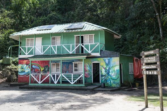 A two-story green house with white railings and vibrant murals on its lower walls, surrounded by dense vegetation. A wooden sign nearby indicates it is located in Los Haitises National Park.