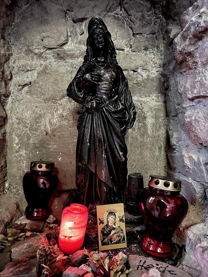 This image features a small, devotional alcove with a religious statue, likely of a saint or similar figure, carved in a dark material and standing prominently. Around the statue are several votive elements: two red candle lanterns, another candle glowing in a clear red holder, and a small religious icon depicting a saint holding a child. The alcove is lined with roughly textured stone walls and the floor is scattered with dried flowers, giving the space a reverent and historical atmosphere. There is a handwritten note partially visible, suggesting a personal significance or a prayer intention linked to this setup.
