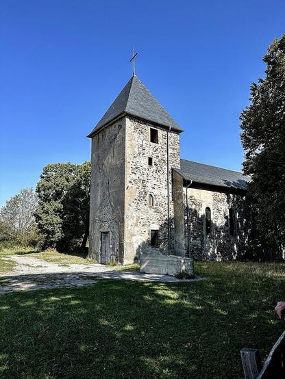 The image shows an old stone church with a tower. The church features a steeply pitched roof and a cross at the top of the tower. The architecture suggests it could be historical, possibly medieval, given the stonework and design. The surroundings are grassy with a clear blue sky above, indicating a peaceful, rural environment. There's a path leading up to the church, enhancing its quaint and serene setting.