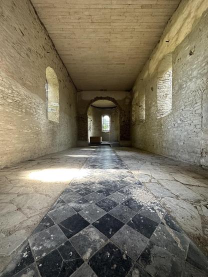 This image shows the interior of a historic building with architectural elements suggesting it could be a church or another type of old religious or communal gathering place. The room features a vaulted passageway and arched windows, which are characteristic of Gothic or medieval architectural styles. The walls are made of rough stone masonry, adding to the antique aesthetic of the space.

The floor boasts an eye-catching checkered pattern made from large, square stone tiles in alternating shades, leading the viewer's eye towards the end of the corridor. The checkered design brings a stark, patterned contrast to the much rougher texture of the walls and the ceiling. Light streams in from the windows, highlighting the textures of the stone and creating a serene atmosphere. Overall, the image exudes a sense of historical atmosphere and peaceful solitude.
