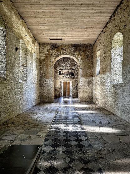 This image shows an interior view of what appears to be a historical building, possibly part of a castle or an old fortress. The corridor is characterized by its stone walls and arches with several windows, creating a sense of antiquity and architectural design common in medieval or ancient buildings. The ceiling is finished with wooden planks, suggesting possibly restored or preserved elements different from the original stone construction. The floor features a striking black and white checkered pattern that adds a refined contrast to the rough texture of the stone walls. The space is illuminated by natural light, which enhances the texture of the stonework and gives the space an airy, open feel. At the end of the corridor, there's a wooden door, indicating access to another part of the structure.