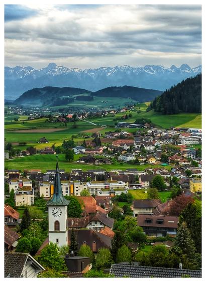 A small village from an elevated viewpoint. The tower of a church clearly stands out. Hills and mountains in the background.