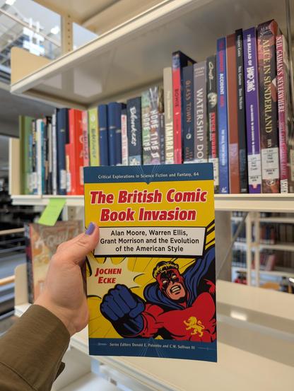 Holding Jochen Ecke's monograph "The British Comic Book Invasion" in front of a bookshelf filled with comic books