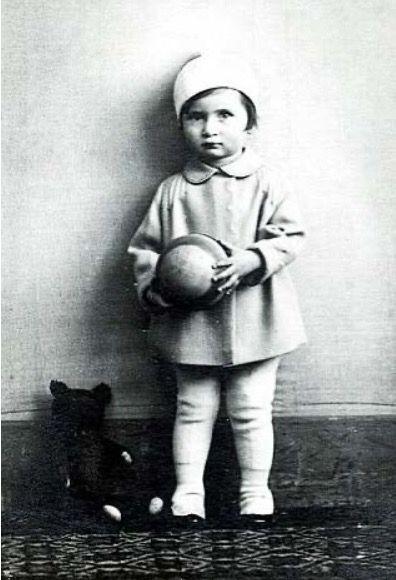 A girl standing against the wall. She is wearing a short jacket and a hat. A toy dog is by her leg. She is holding a round object in her hands.