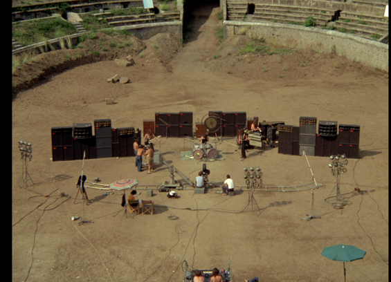 Scene fra Pink Floyd at Pompeii.
Bandet står i amfiteatereret med deres højtalere og instrumenter. Der er intet lysshow og intet publikum. Kun et par teknikere og kamerafolk. Der ligger dolly tracks foran dem og der står et kamera på en lille kran. Det må være en varm dag, for flere af den har bar overkrop