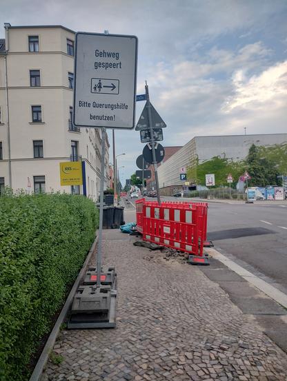 Fußweg neben einer Straße mit Mehrfamilienhäusern und Gewerbe. Im Hintergrund, halb auf dem Fußweg, stehen Bauzäune. Im Vordergrund ein Straßenschild mit der Aufschrift "Gehweg gespeert [sic] - Bitte Querungshilfe benutzen!"