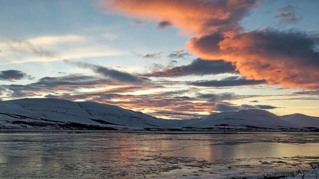 A colour photo of a sunrise. The foreground is a frozen expanse of water and on the horizon is a line of snow-covered mountains. The sky is clear apart from streaks of cloud above the mountains and a block of low cloud in ter right. All of the clouds are tinted orange by the rising sun, and this is reflected in the surface of the ice.