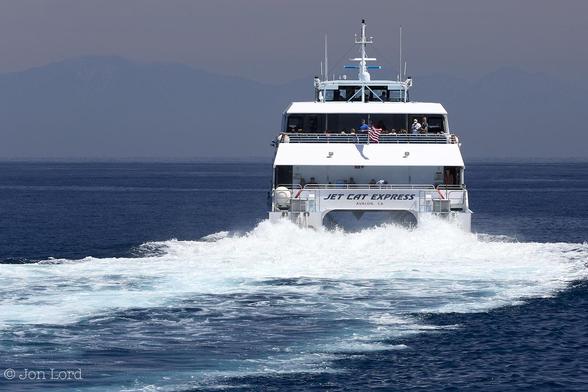 This Is A Colour Seascape Photo In Landscape Format. Covering The Lower Two Thirds Is An Expanse Of Dark Blue Calm Sea. To The Right Of Centre Is The Stern Of A SeaCat Fast Ferry Under Way.  The View Of The SeaCat Is Aligned So None Of The Sides Are In View, Just The Stern. Aft Of The Ferry Is A Mix Of Blue Ocean And White Froth From The Powerful Water Jet Nozzles. The Ship Being A Catamaran Has Two Parallel Hulls Separated By Five Or So Metres With Two Main Decks And A Bridge Above The Upper Deck. The Aft Section Of Both Decks Are Open And Passengers Can Be Seen. Some Distance Ahead Of The Ferry Is A Tall Mountainous Ridge That Stretches From The Left To The Right Margins. On The Left Side A Little Late Spring Snow Can Just Be Made Out. Above The Sky Has A Slight And Very Light Orange Hazy Tint To It. 
The Location: Santa Catalina, California. The Body Of Water Is The San Pedro Channel (The Pacific) And The Mountain Range Is The San Gabriel Mountains. The Ferry Is The 'Jet Cat Express' Bound For Long Beach (2015).
