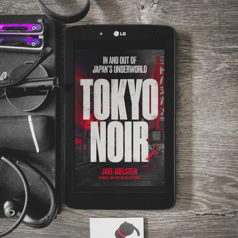 in and out of japan's underworld, tokyo noir by jake adelstein book cover, white letters on black, grey, and red city street, with signs and archtiecture unique to japan. black framed LG tablet is placed on a dark grey wooden table, with various dark items strewn around, such as round shades, lighter, earphones, bracelet, notebook.