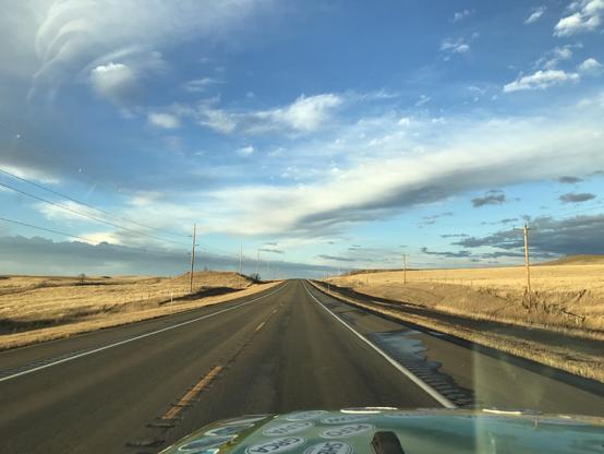 A two-lane stretch of pavement with dried, golden grasses on either side, and high clouds overhead