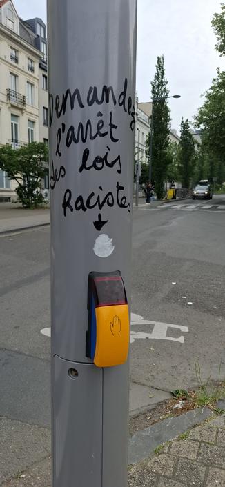 Sur un poteau gris on voit un boîtier jaune et noir avec une main dessinée dessus, c'est un bouton pour demander le feu piéton.
Sur le poteau il y a un message écrit "demandez l'arrêt des lois racistes".