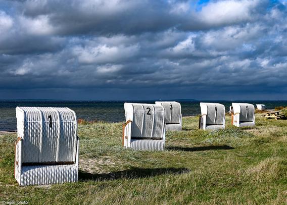 Blick von hinten auf fünf weiße große Strandkörbe, die auf einer Wiese direkt an der Ostsee in Richtung Wasser stehen.
Der Himmel ist stark bewölkt, die tiefstehende Sonne läßt die Körbe lange Schatten werfen. /
View from behind of five large white beach chairs standing on a meadow directly on the Baltic Sea, facing the water.
The sky is heavily overcast, and the low sun casts long shadows on the chairs.